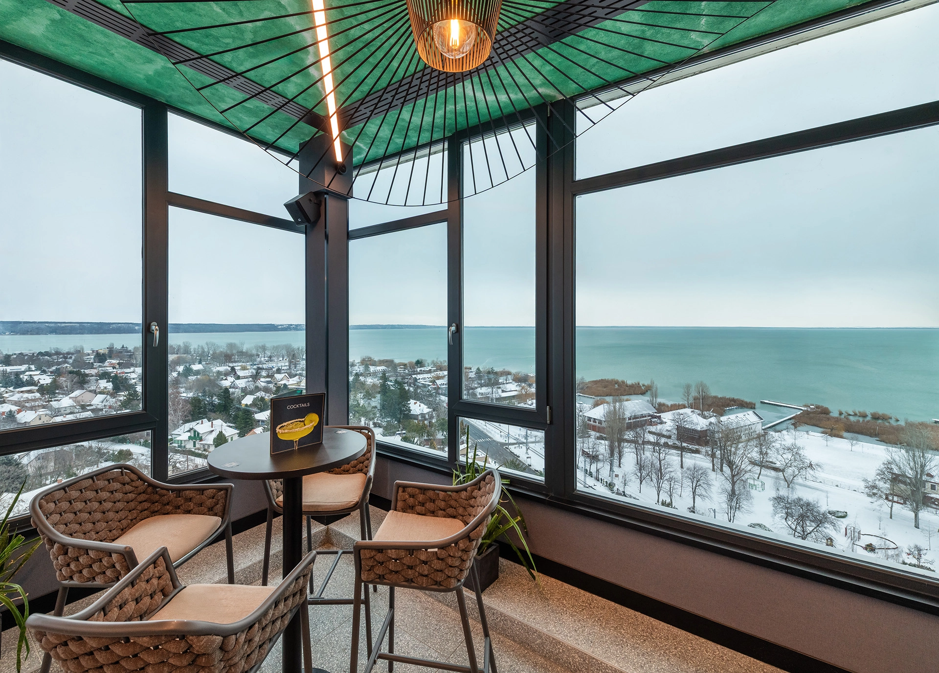 Hotel rooftop lounge interior with panoramic windows overlooking a snowy lakeside town and frozen landscape