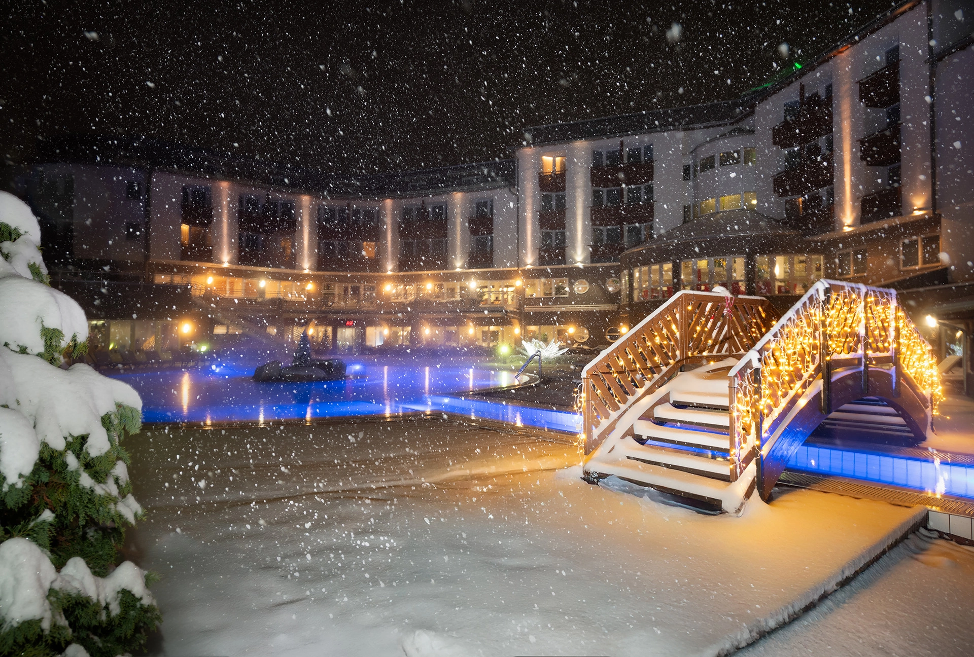 Outdoor thermal pool and wooden bridge at night during heavy snowfall with warm lights and illuminated hotel facade