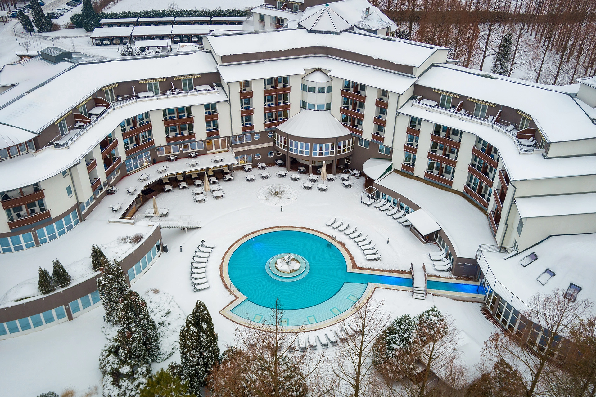 Aerial drone view of a snow-covered hotel courtyard with circular thermal pool and surrounding resort buildings in winter