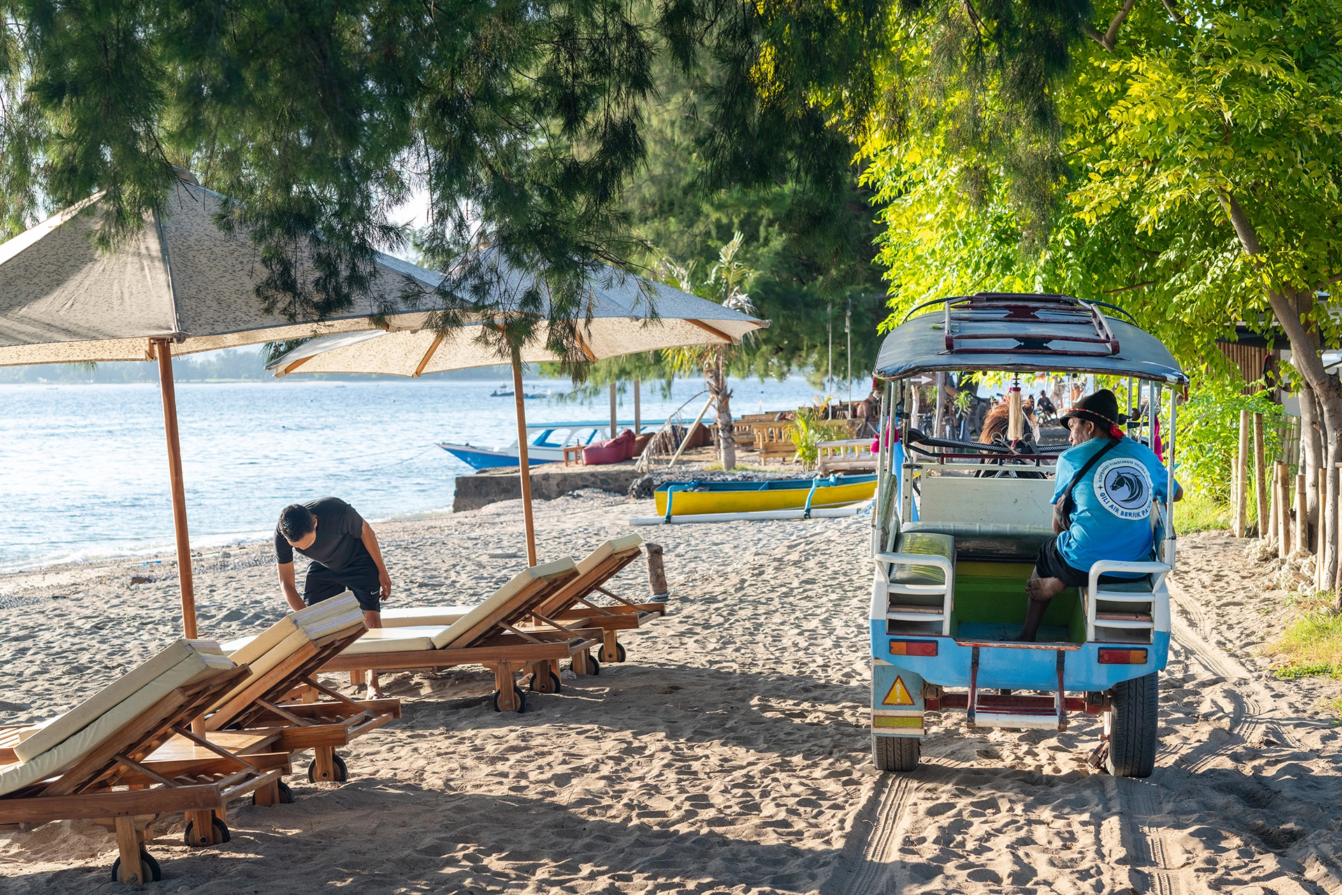 Beach loungers and a traditional island cart under tropical trees on Gili Air beach at sunrise.